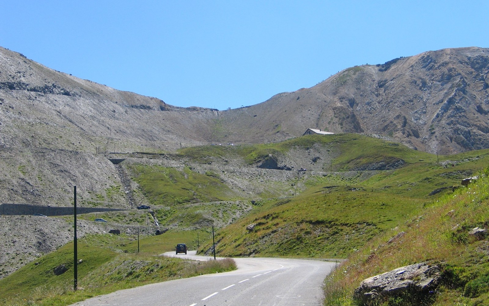 A winding mountain road climbs through grassy slopes toward the barren peaks of the Galibier