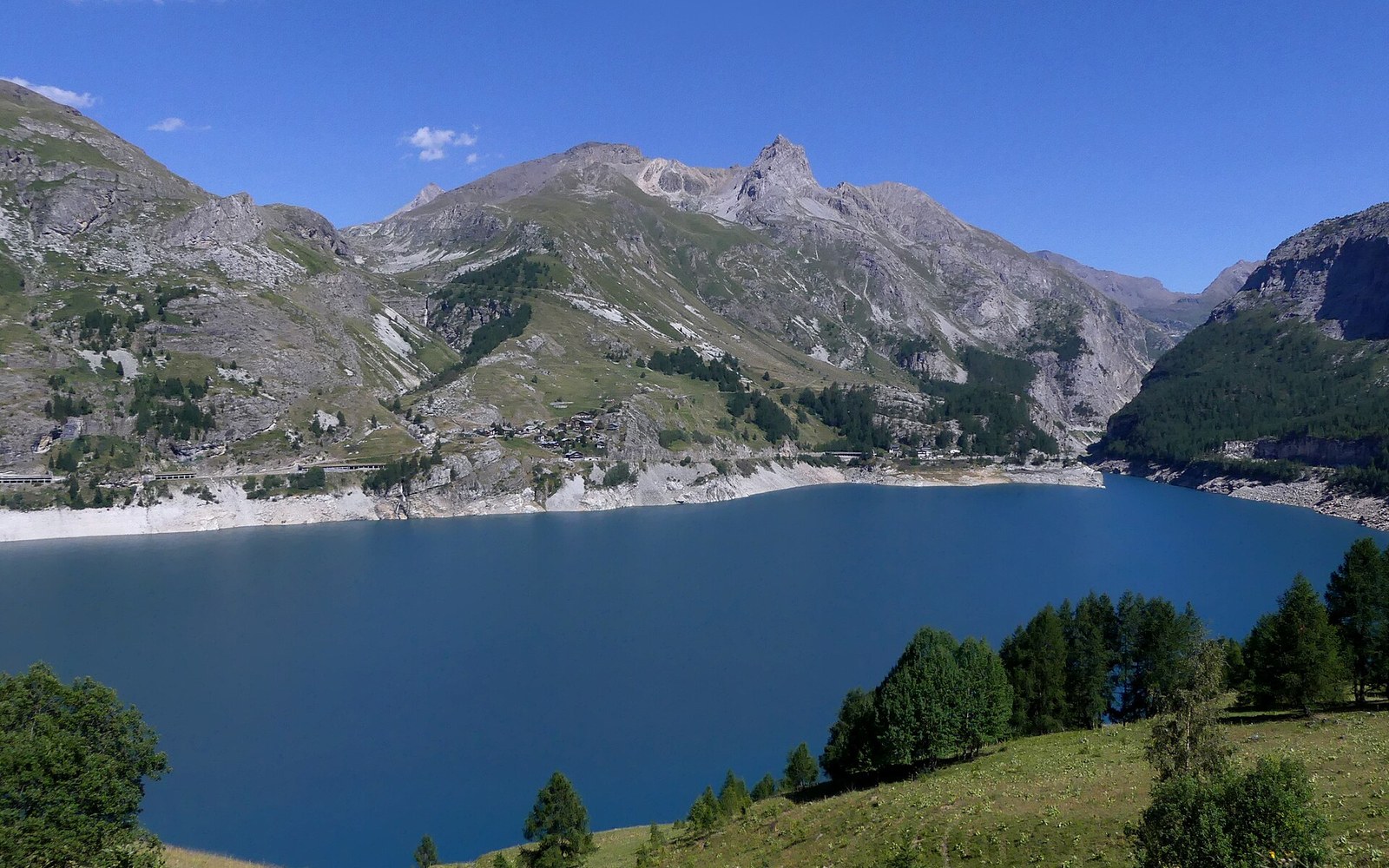 Turquoise reservoir surrounded by steep alpine peaks and green slopes under a clear sky